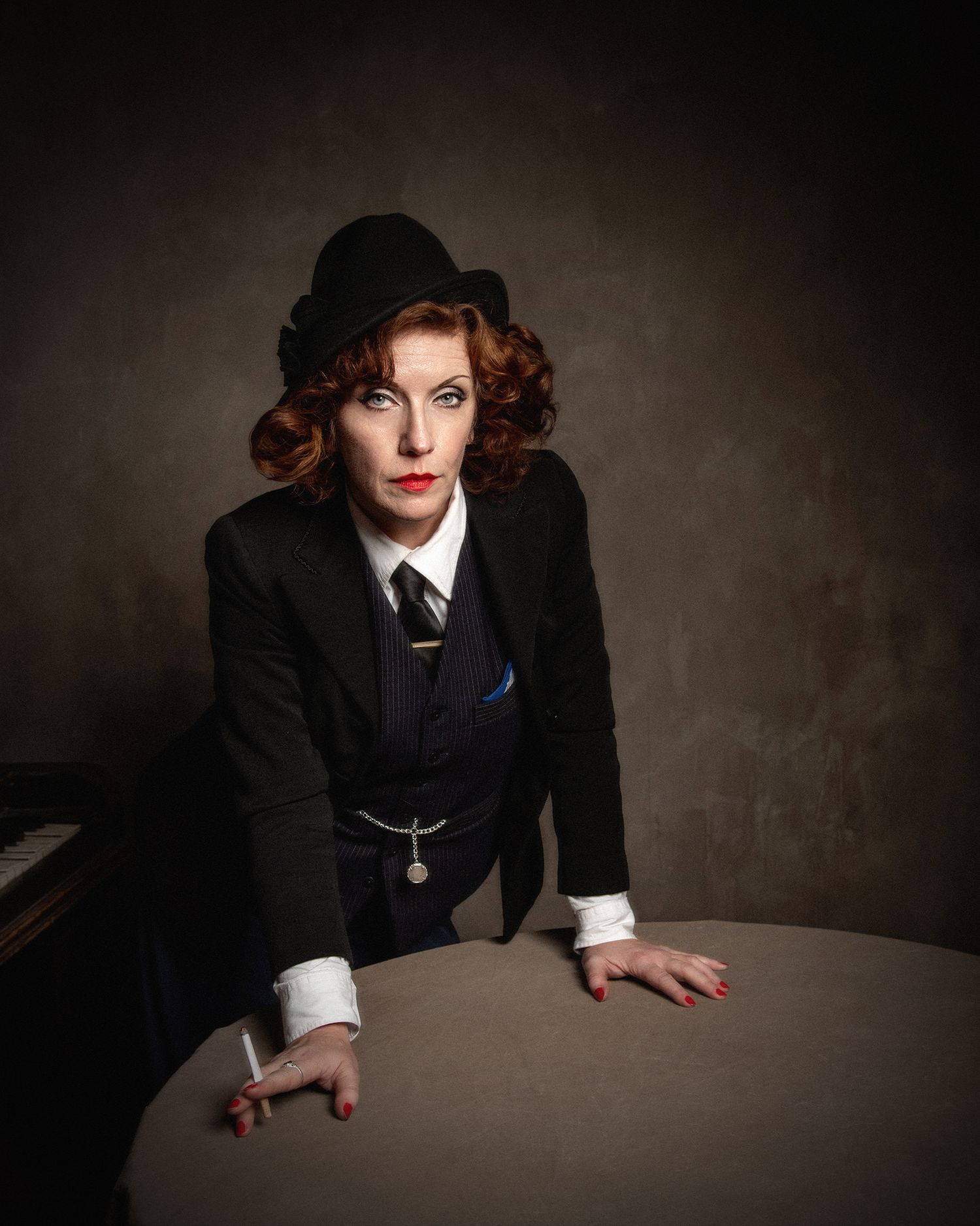 Dramatic portrait of figure in 1920s masculine-style suit and hat leaning over table.