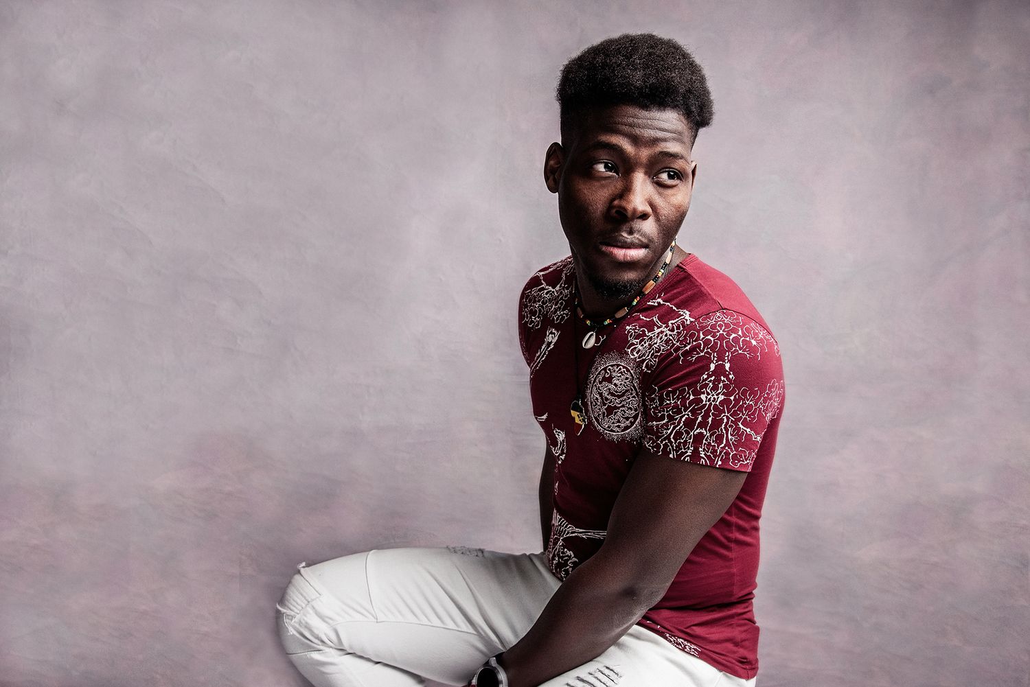 Studio portrait of person wearing burgundy patterned shirt and white pants against purple background.