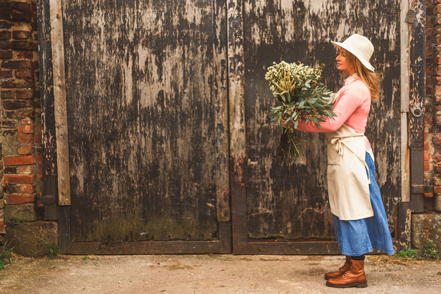 Someone in a sunhat and apron holds a bouquet of wildflowers against a rustic weathered wooden door background.