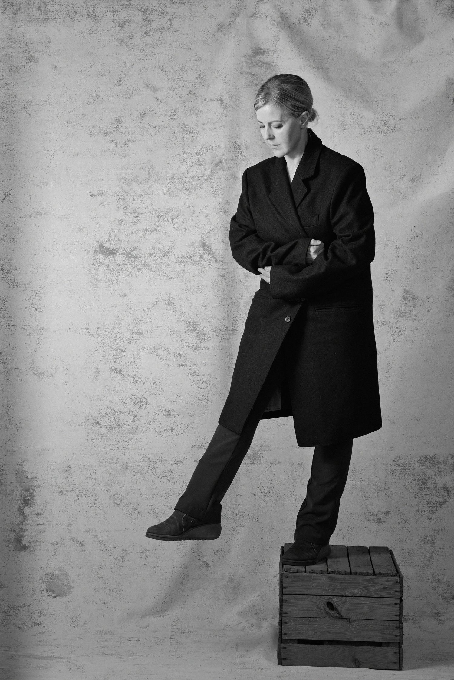 Black and white portrait of a woman in oversized black coat standing with one foot on a wooden crate.