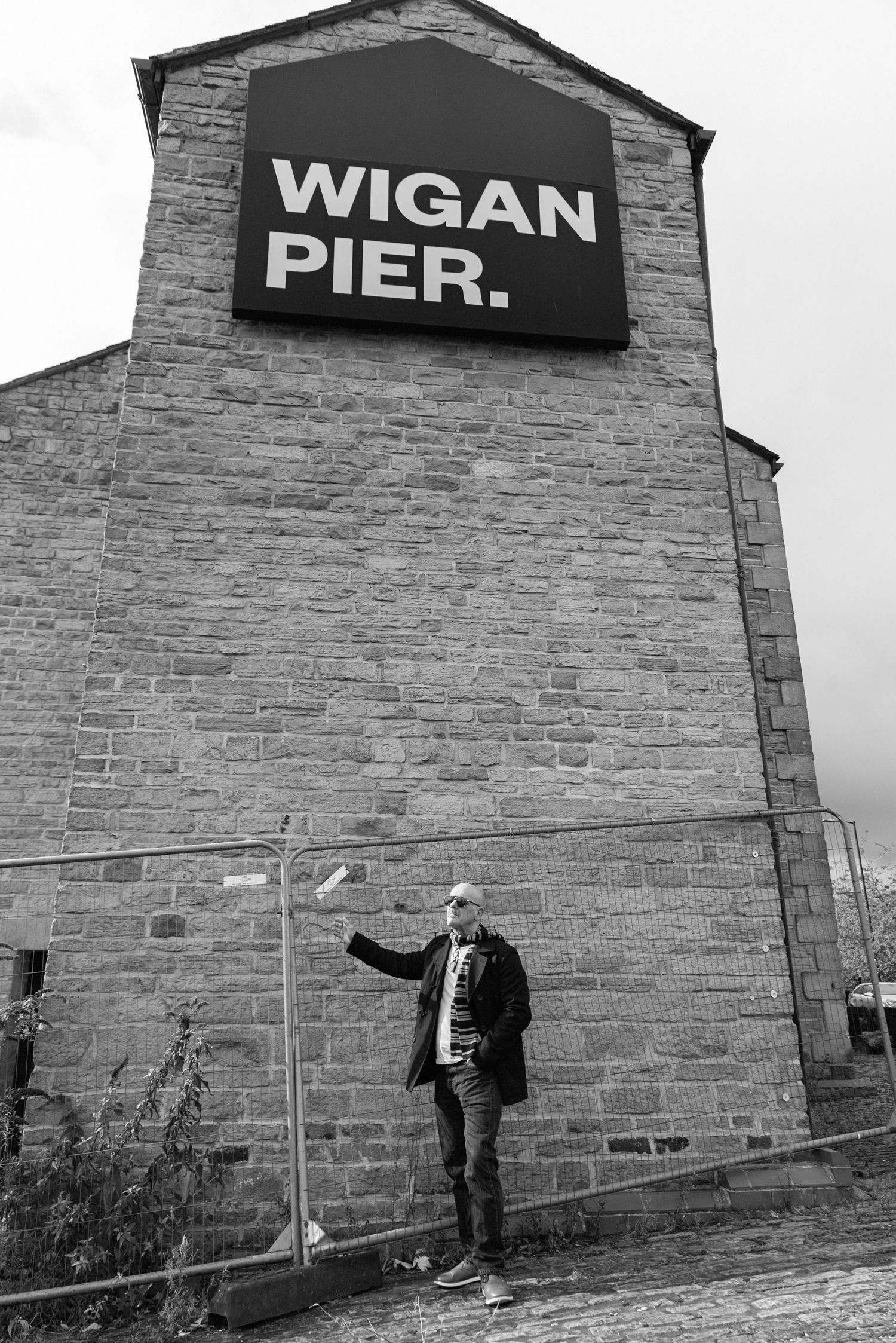 Black and white photo of a person pointing at Wigan Pier sign on a historic brick building in Wigan, England.