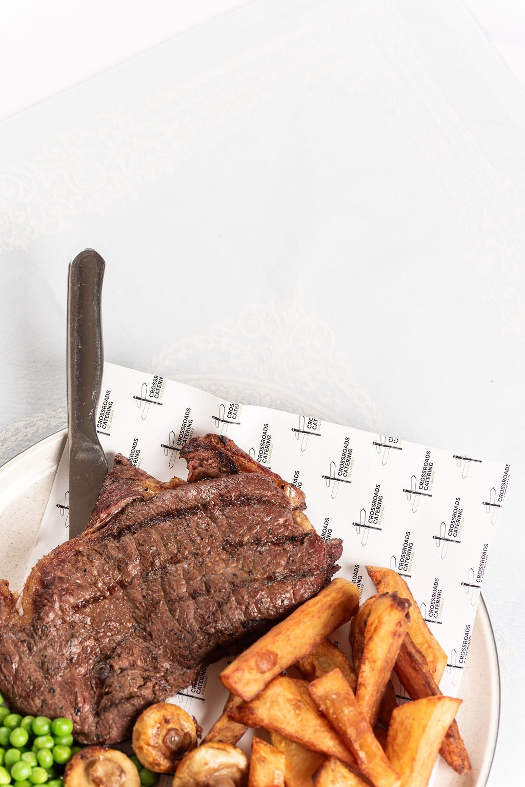 Grilled steak with chunky chips, peas, and mushrooms served on a white plate with a knife.