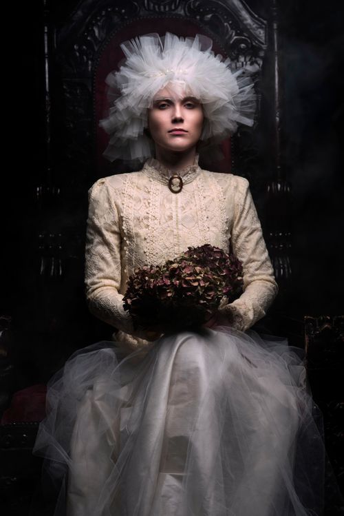 Woman in vintage white lace gown and tulle headpiece sits on dark throne holding dried flower bouquet.