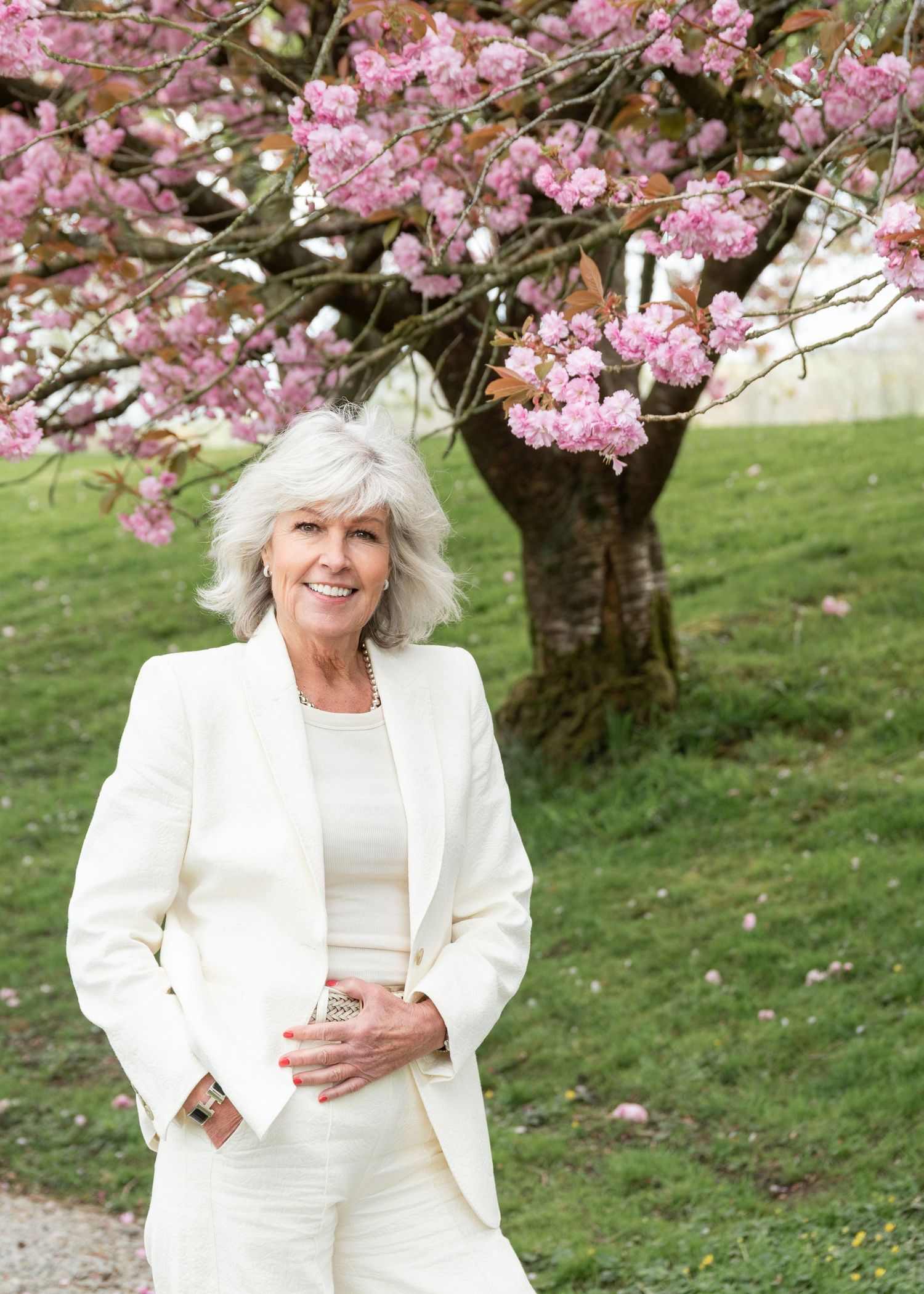 Elegant woman in white suit smiling under pink cherry blossom tree on green lawn in spring.