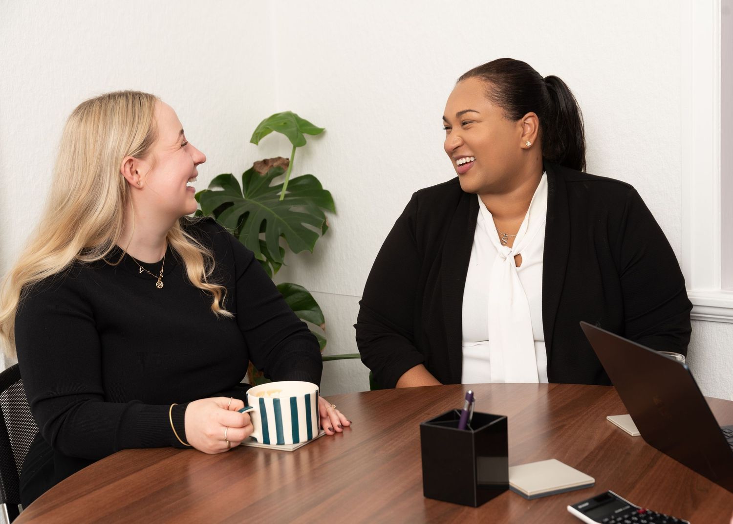Two women smiling and laughing together at a office table, one holding a striped mug, with a laptop nearby.
