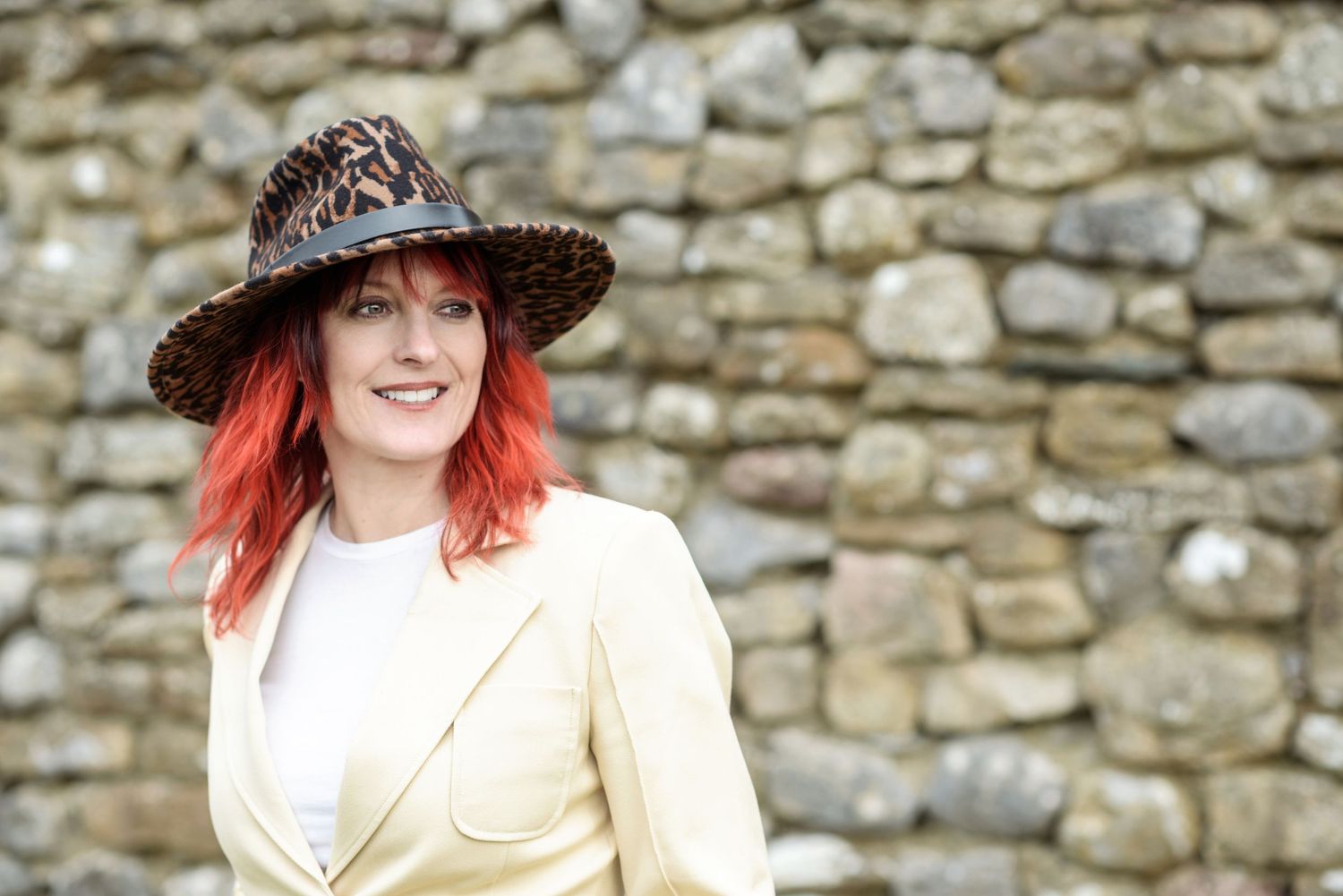 Woman with red hair wearing a leopard print hat and yellow blazer smiling against a stone wall.
