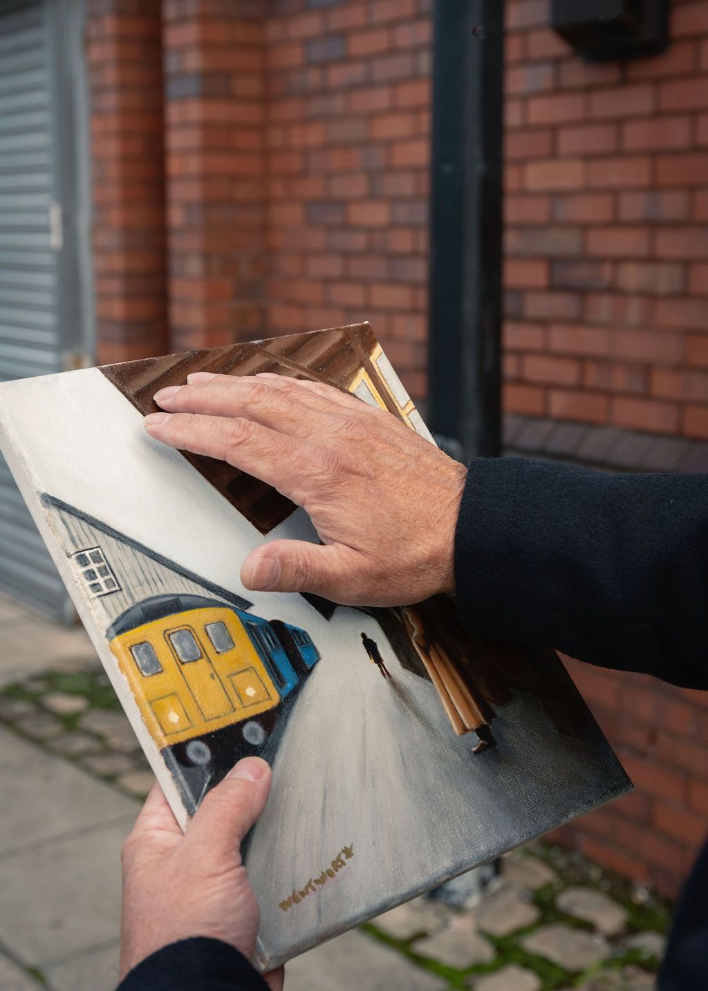 A man's hands holding a small oil painting of a yellow train at a station, displayed outdoors against a brick wall backdrop.