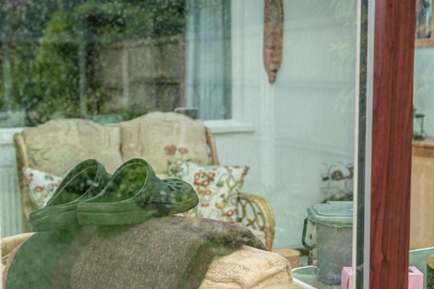 Cozy conservatory interior viewed through glass, featuring wicker furniture, green throws, and cushions with garden view.