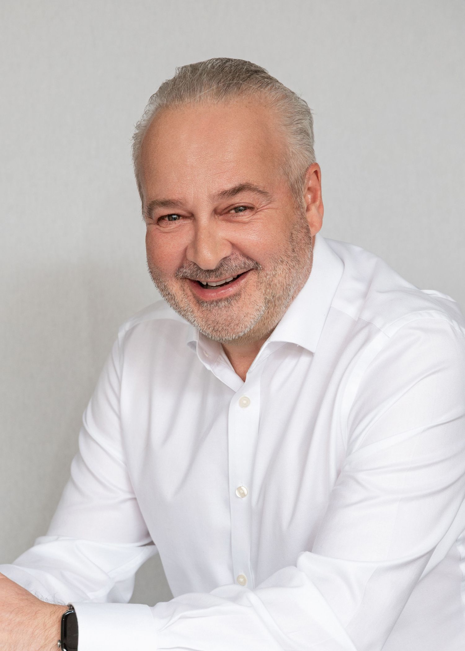 Smiling middle-aged man with gray hair and beard wearing a white shirt against a light background.