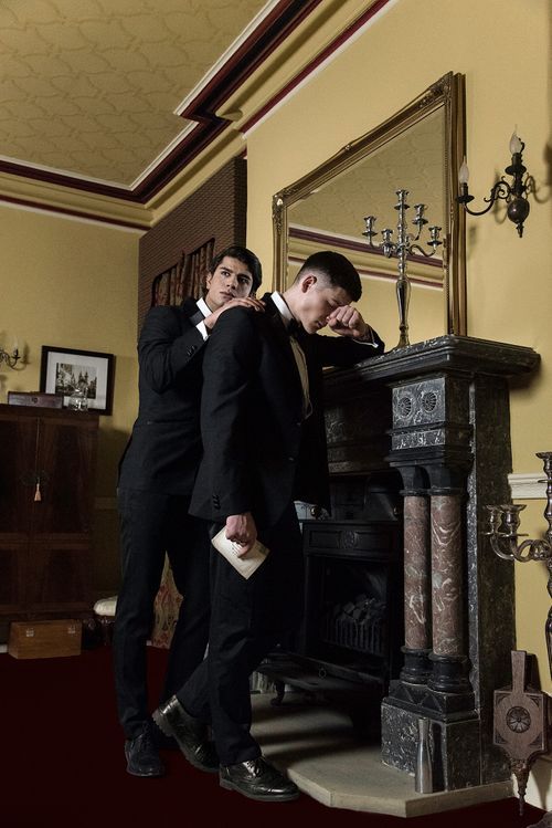 Two men in black suits pose dramatically by an ornate fireplace in an elegant Victorian-style room with gold mirror.