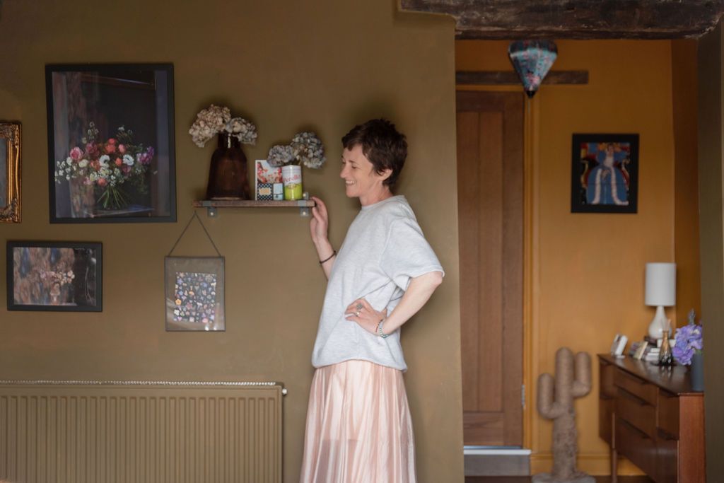 Elderly woman in pink skirt and white top standing by wall with framed artwork and decorative shelf in cozy room.