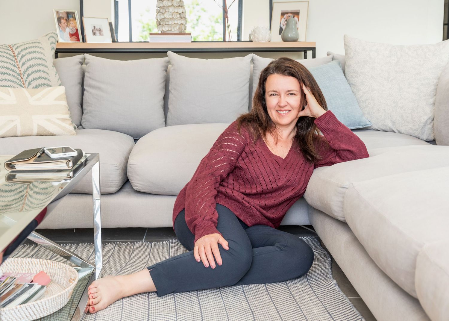 Woman in burgundy sweater and navy leggings sitting on floor in modern living room with gray sectional sofa.
