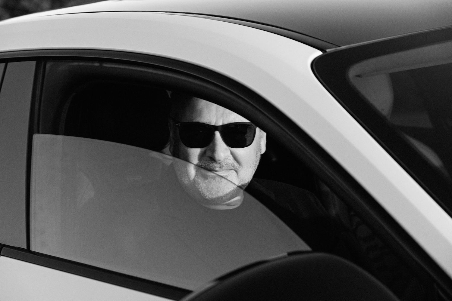 Black and white photo of an older man wearing sunglasses sitting in a white car looking out the window.