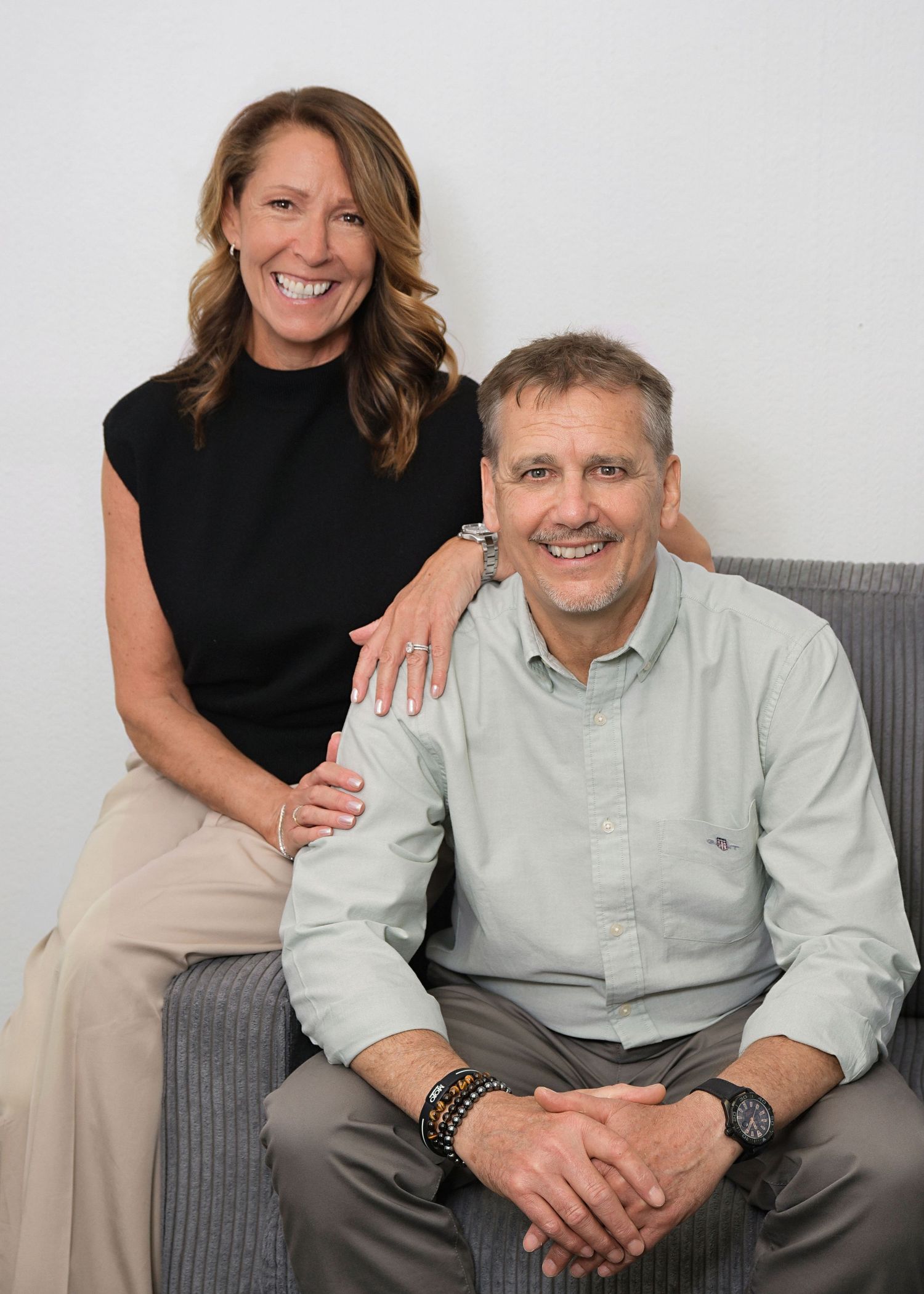 A smiling man and woman pose together on a gray couch, wearing casual business attire in a bright, modern setting.