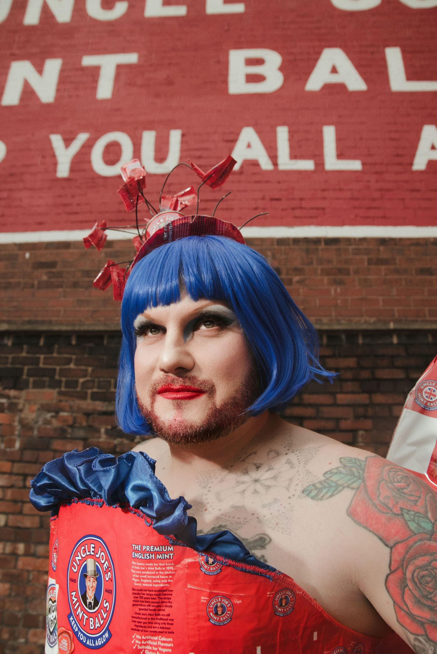 Person in blue wig, red dress, and floral headpiece poses outdoors near a red brick wall with signage.