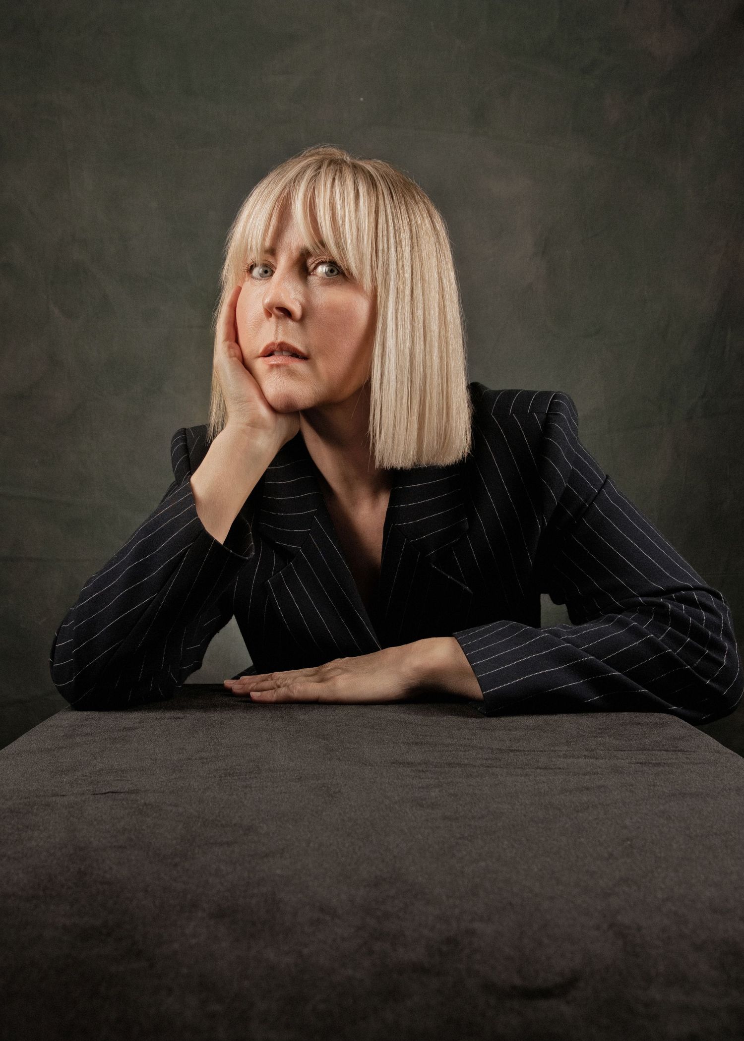 Blonde woman in pinstripe blazer rests chin on hand, gazing upward thoughtfully while seated at a table.