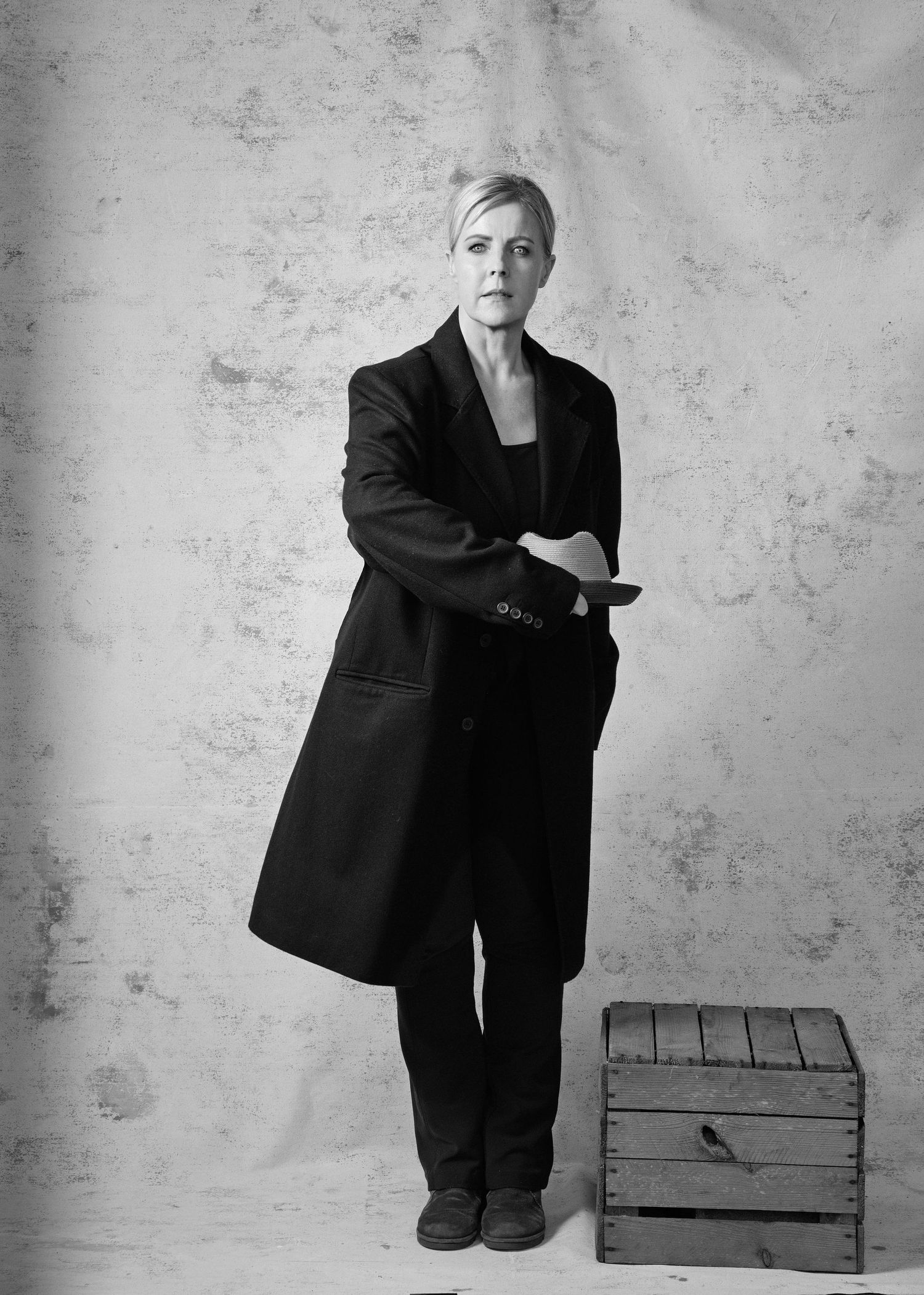 Black and white portrait of a woman in a long dark coat standing beside a wooden crate.