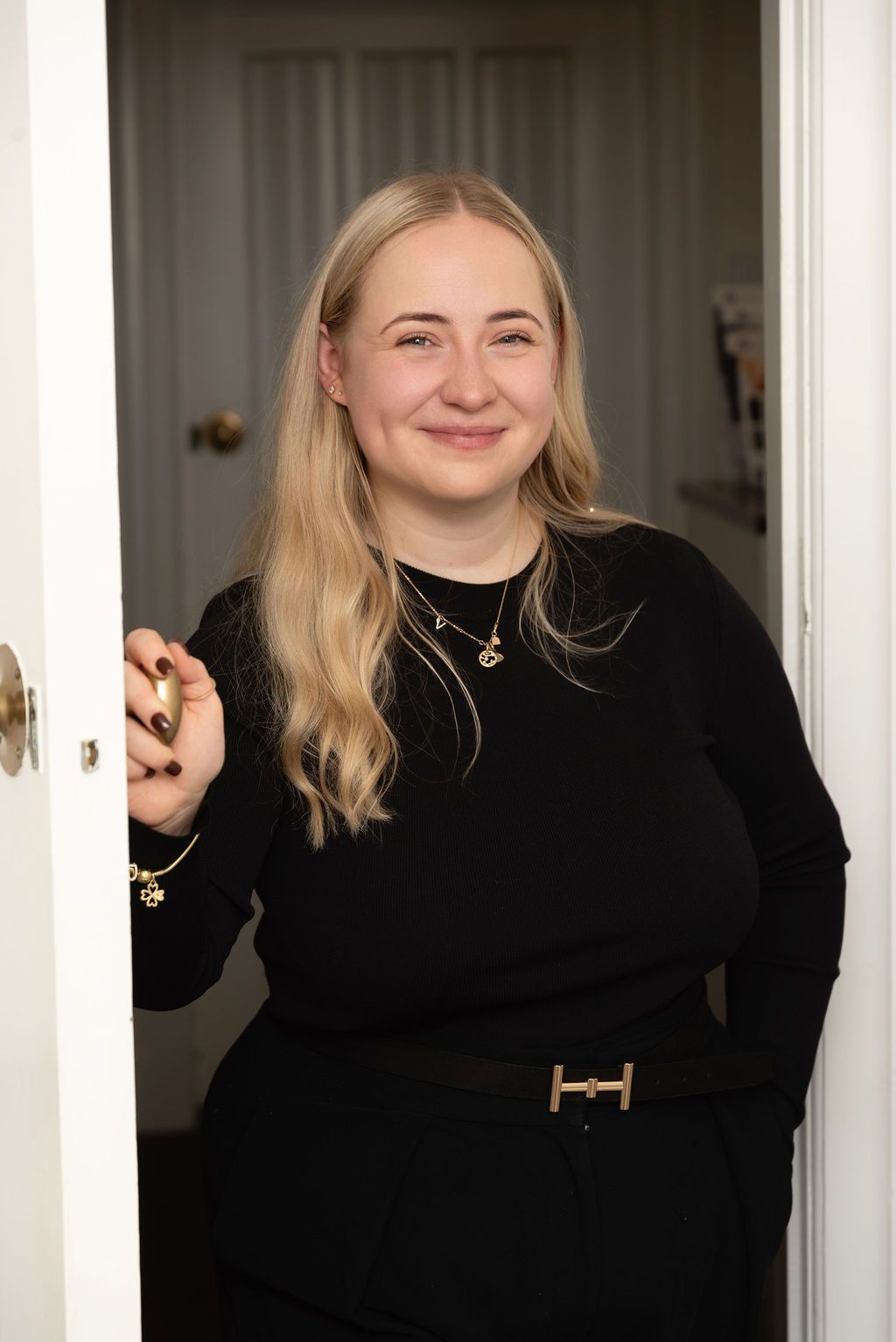 A person wearing a black sweater and gold belt stands smiling in a doorway with light blonde hair.