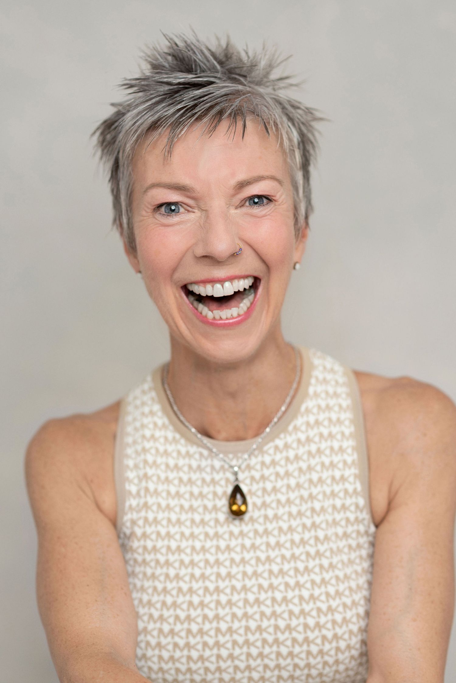 Energetic portrait with spiky grey hair wearing cream knit top with pendant necklace.