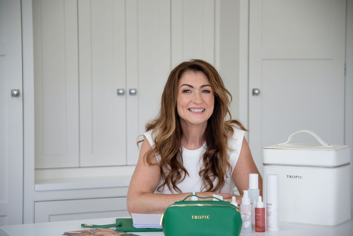 Smiling woman with brown hair sits at a vanity displaying green Tropic skincare products and white storage box.