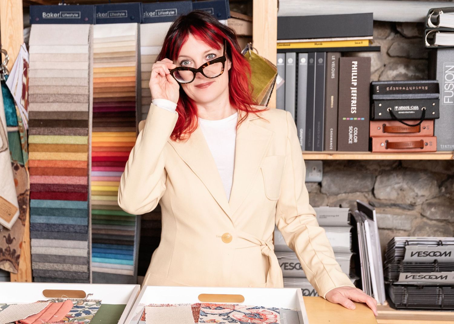 Red-haired woman in beige blazer adjusting glasses in a fabric and design studio with colorful swatches.