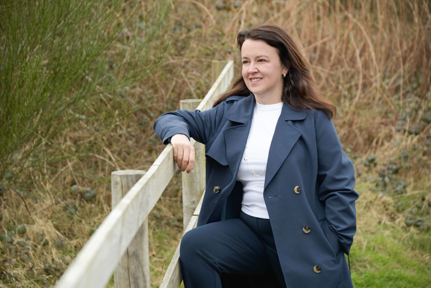 Woman in navy blue coat leaning on wooden fence outdoors, smiling in a natural green landscape setting.