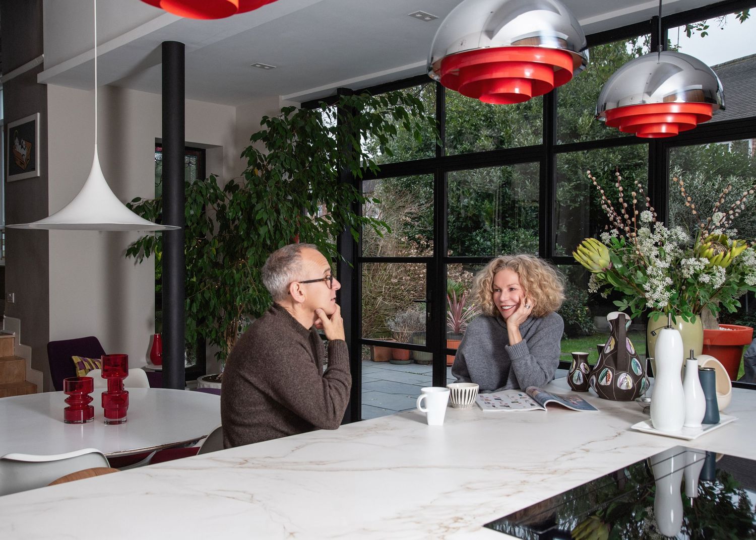 Two people enjoying coffee at a modern white kitchen counter with red pendant lamps and large windows overlooking greenery.