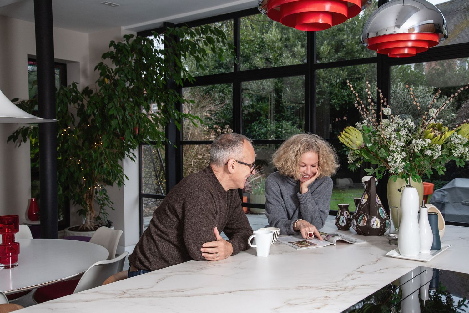 Modern restaurant interior with white tables and red pendant lamps hanging from ceiling near outdoor garden area.