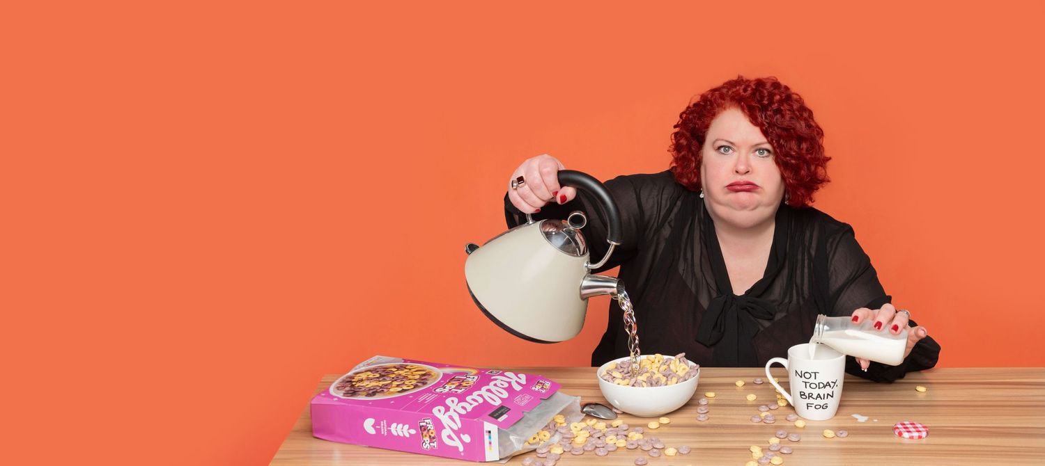 Red-haired woman in a black top pours water from a kettle into a cereal bowl on a wooden table against an orange background.