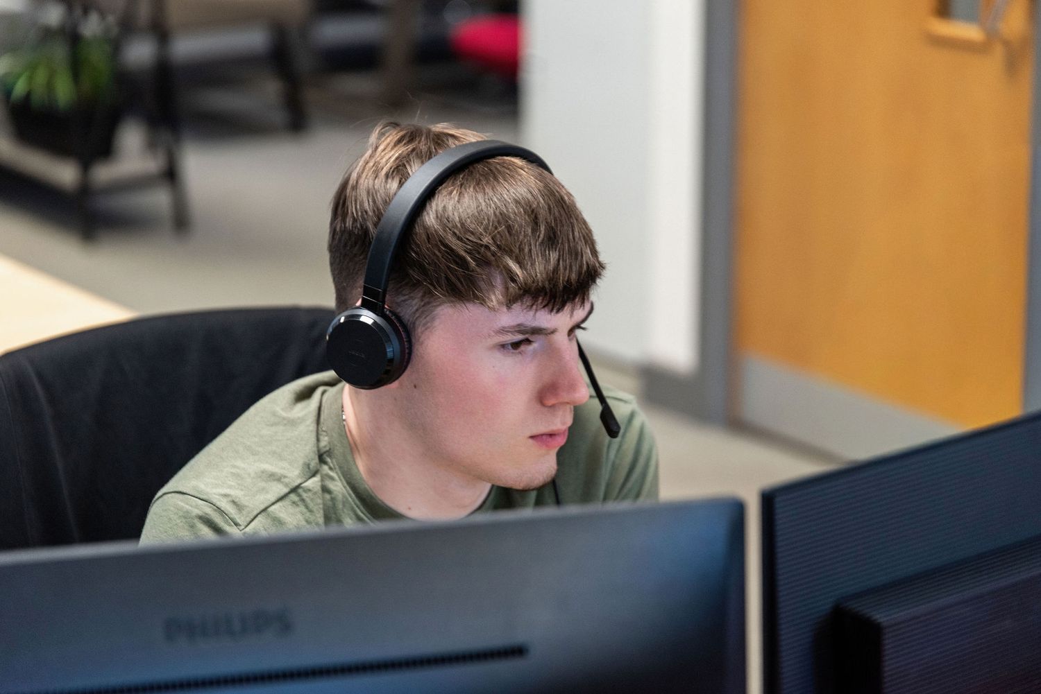 A focused teenage boy wearing headphones works intently at a Philips computer monitor in an office setting.