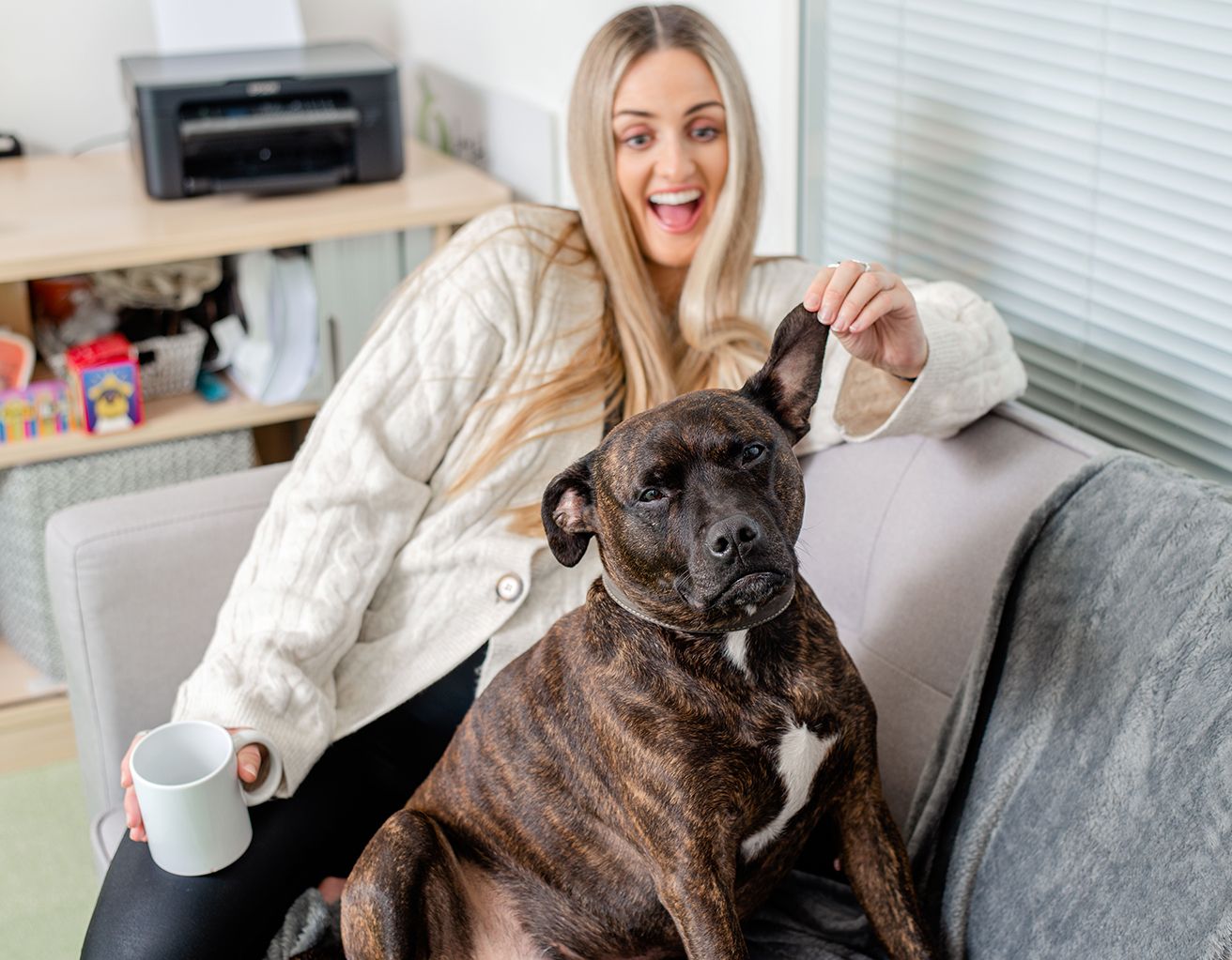 A person sits on a couch with a large dark-colored dog while holding a white coffee mug.