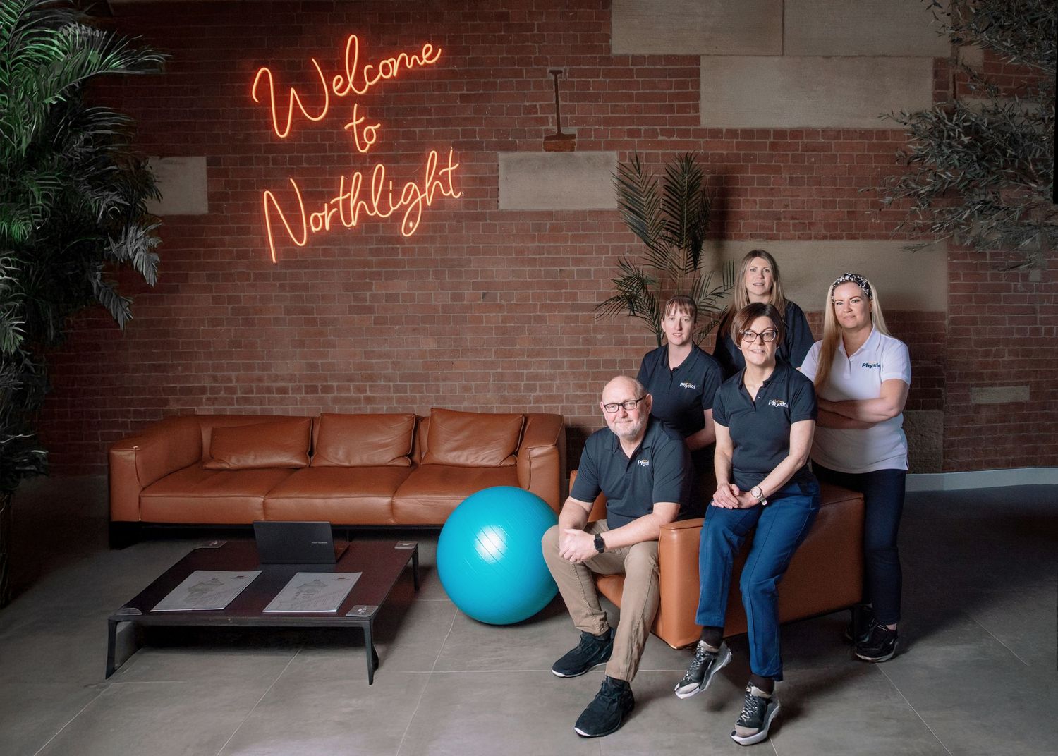 A modern office space with a brown leather couch, neon 'Welcome North Light' sign, and an exercise ball on the floor.