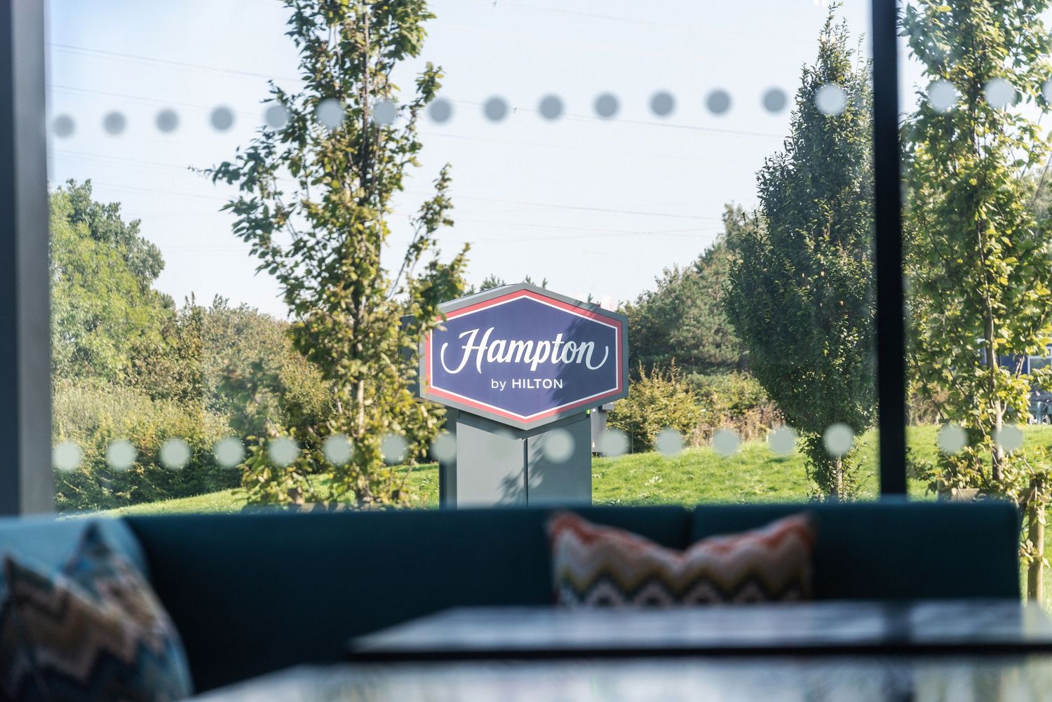 Hampton hotel sign visible through window with outdoor greenery and trees.