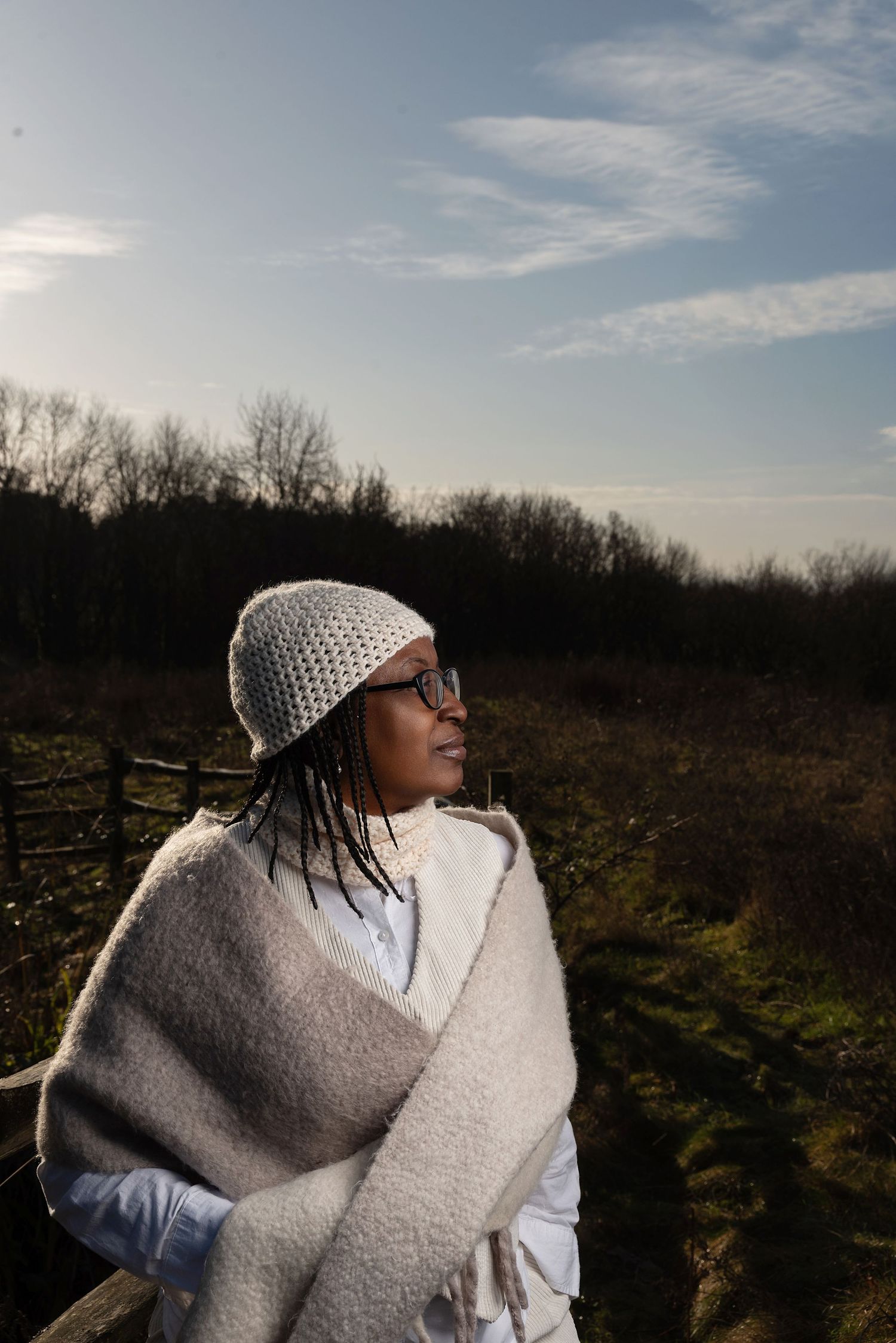 A person wearing a gray beanie and white sweater stands against a natural outdoor backdrop at sunset.