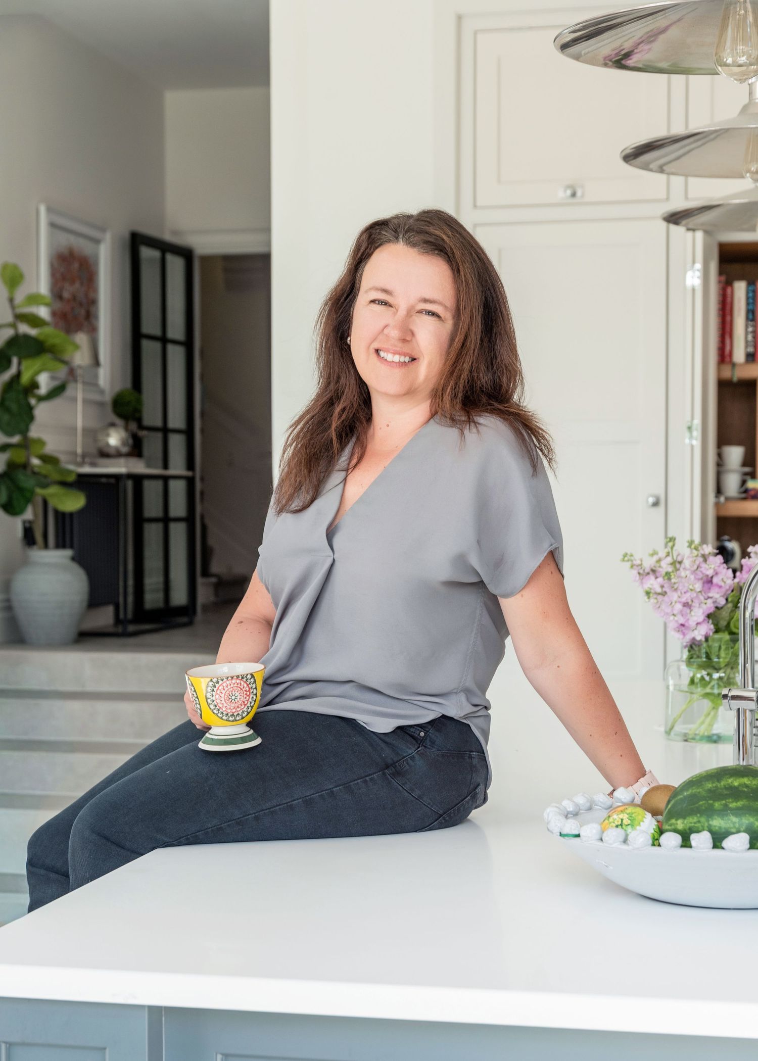 Woman smiling in modern white kitchen, sitting on counter with colorful teacup and fresh fruit nearby.