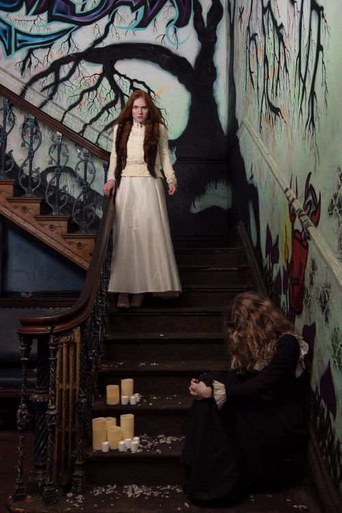 Two women in Victorian dresses on graffiti-covered stairs with candles, creating a gothic, mysterious atmosphere.