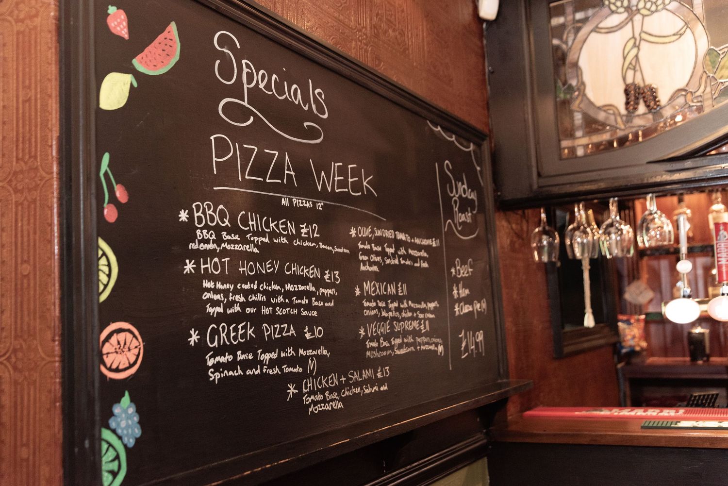 Chalkboard menu displaying Pizza Week specials with colorful toppings illustrations at a cozy restaurant bar.