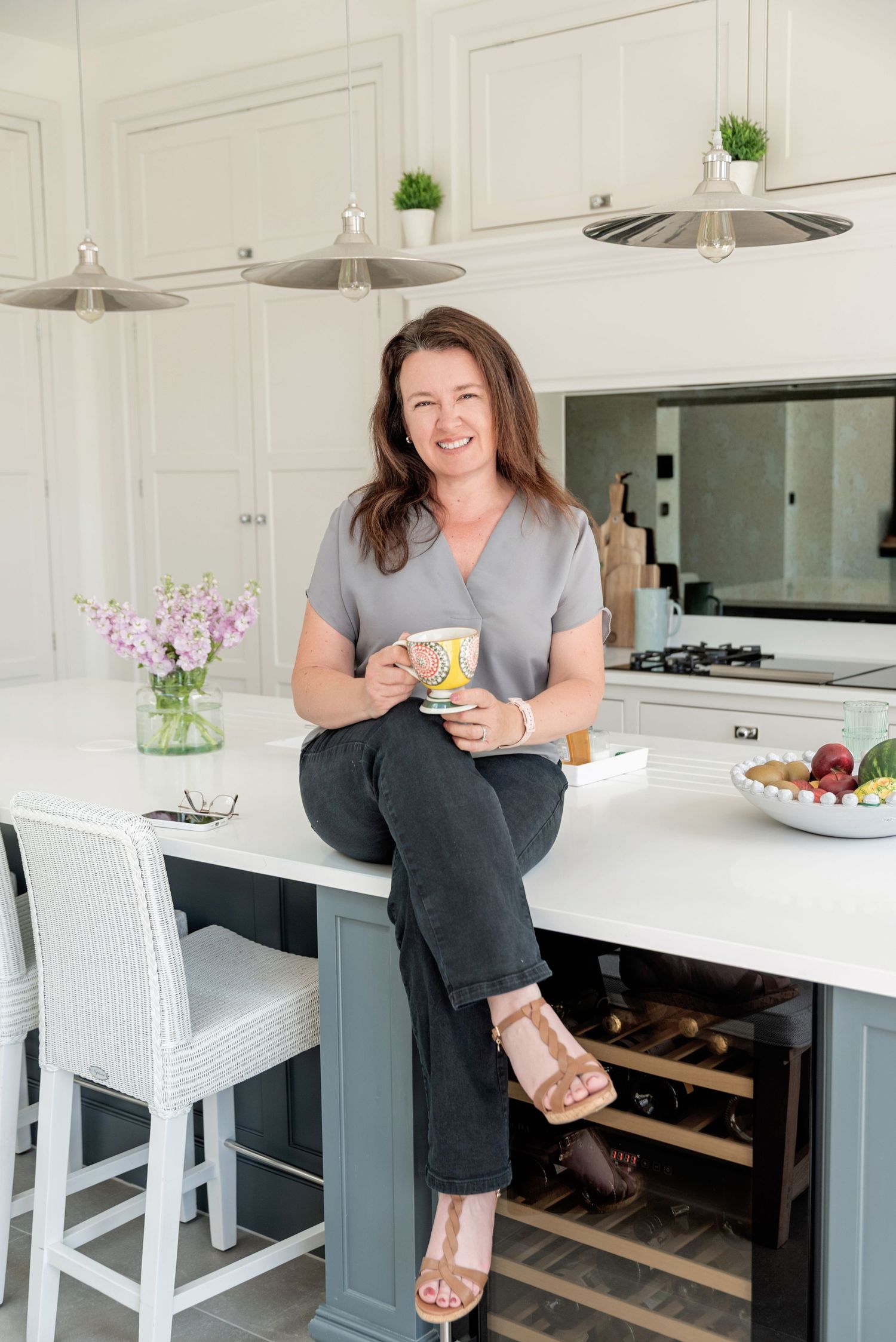 Woman smiling in modern white kitchen, sitting on island counter holding colorful mug, wearing grey top and black jeans.