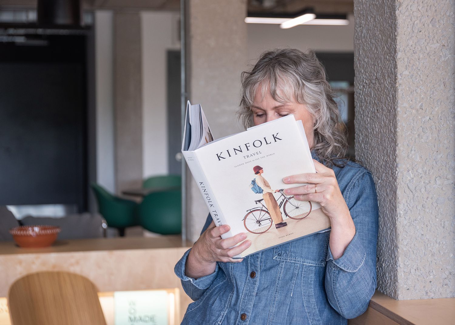 Woman with gray hair reading a Kinfolk magazine, leaning against a wall in a modern cafe setting.