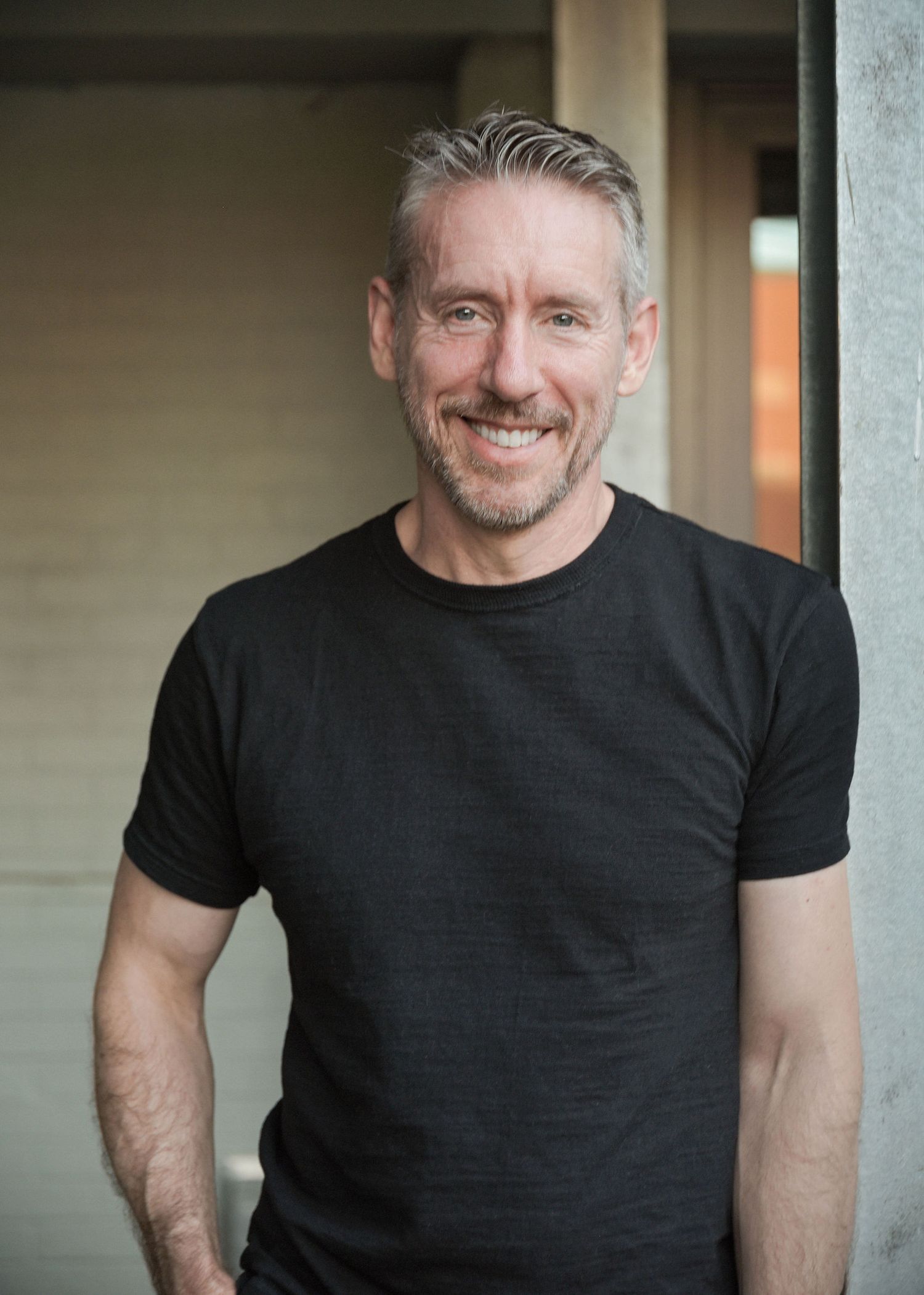 Smiling middle-aged man with gray hair wearing a black t-shirt, posing casually in a modern outdoor setting.