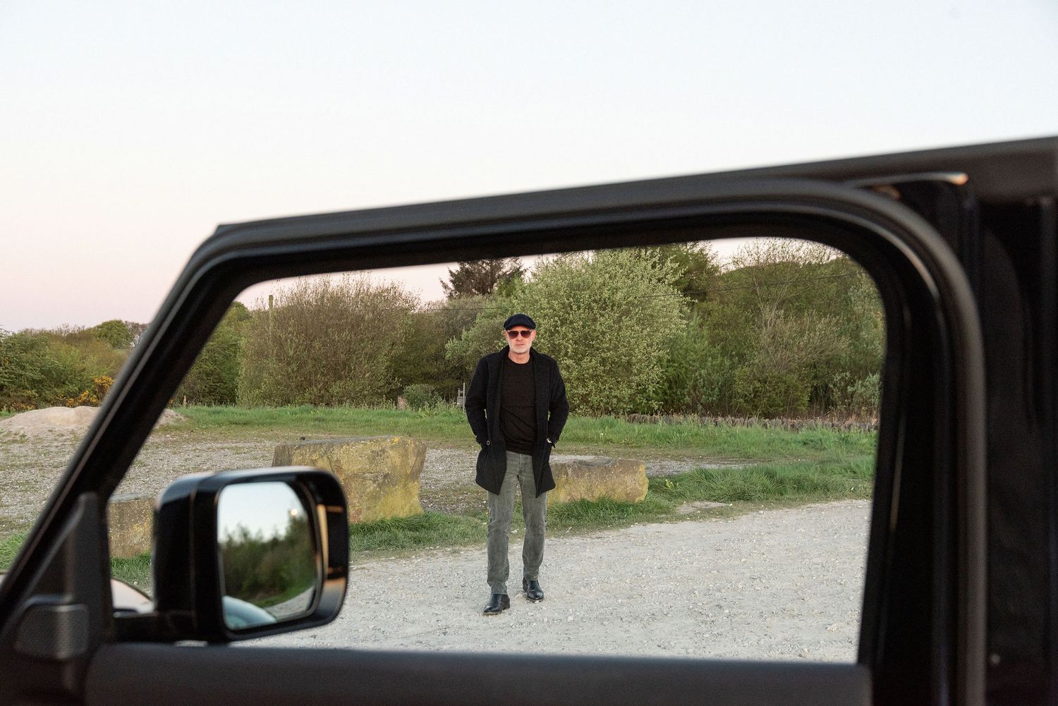Man in black coat and sunglasses standing on gravel path, viewed through open car door window at dusk.