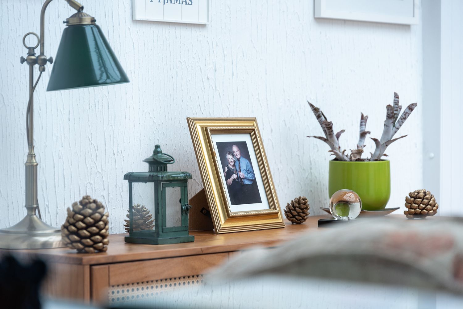 Cozy wooden dresser with gold photo frame, green lantern, pine cones, potted plant with feathers, and vintage desk lamp.