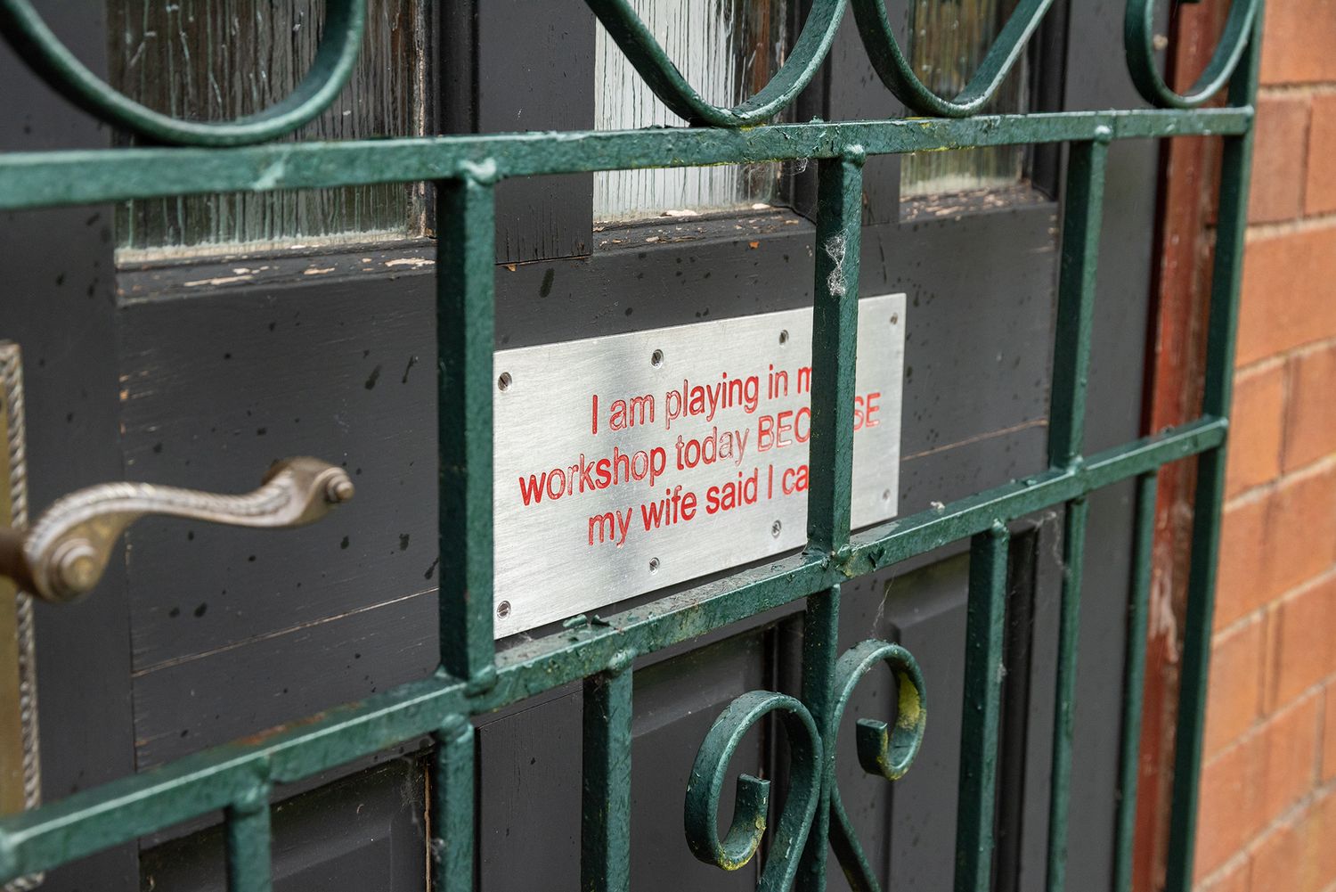 A humorous sign on a green metal gate reads a funny message about playing in the workshop because his wife said he could.