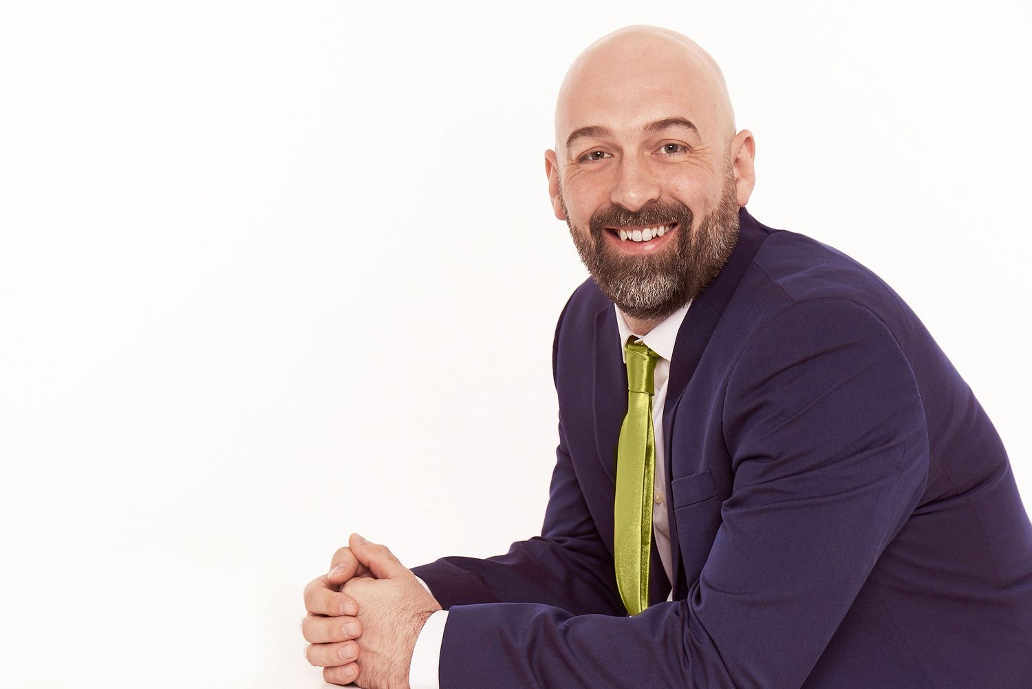 Professional in dark suit and lime tie sits with clasped hands against white background.