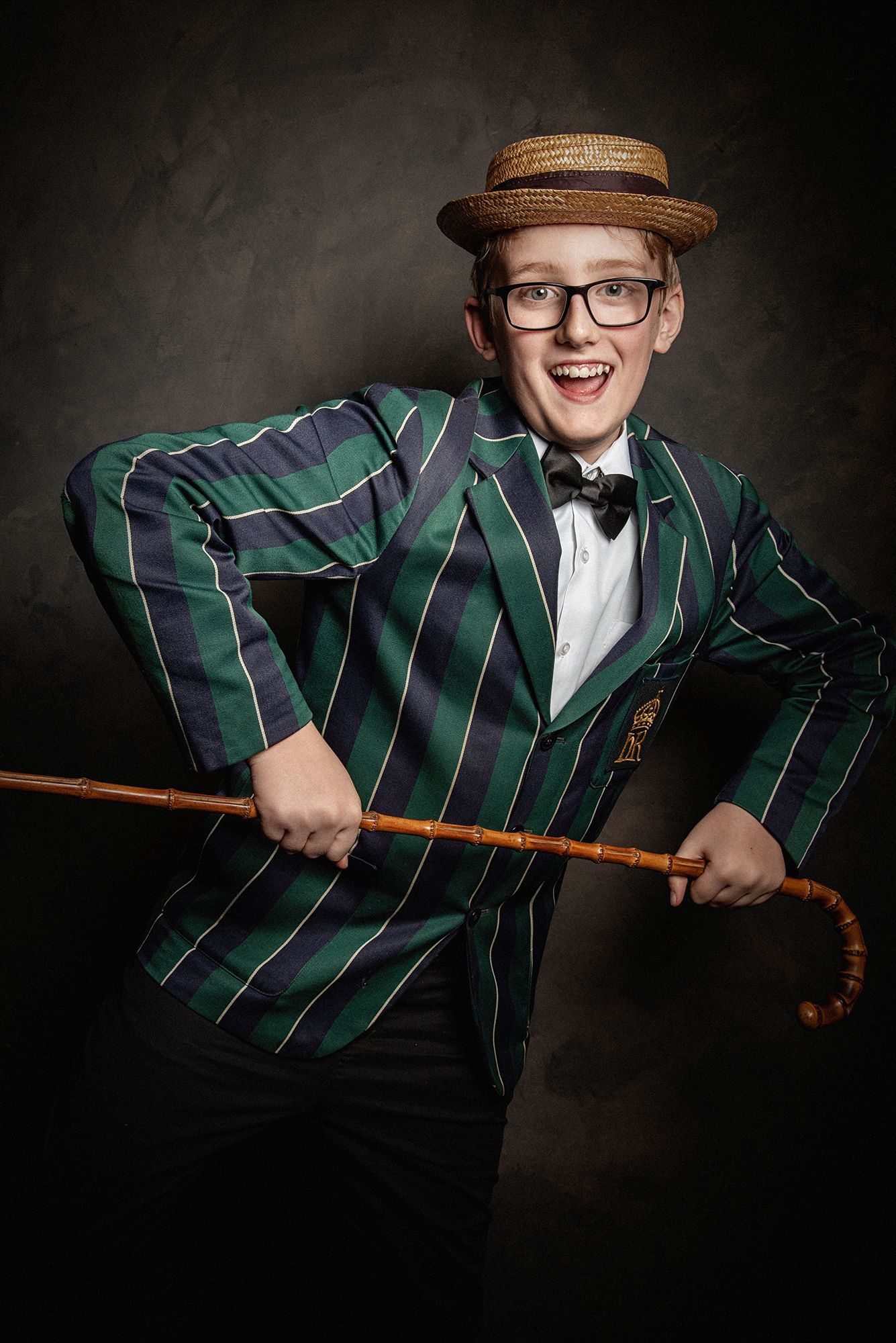 A whimsical portrait featuring a green striped boater jacket, straw hat, bow tie, and walking stick against a dark background.