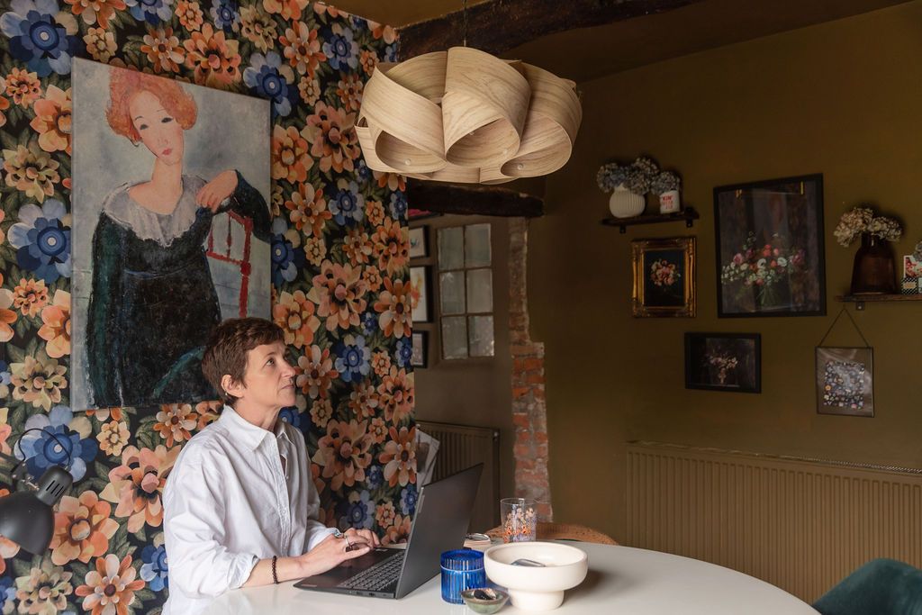 Man working on laptop at white table in eclectic room with floral wallpaper and portrait painting.