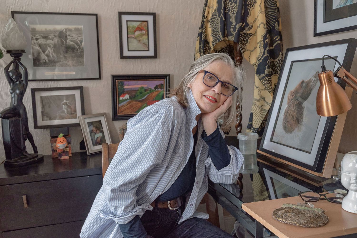 Woman with gray hair and glasses smiles at desk surrounded by framed artwork and eclectic decor in a cozy studio.