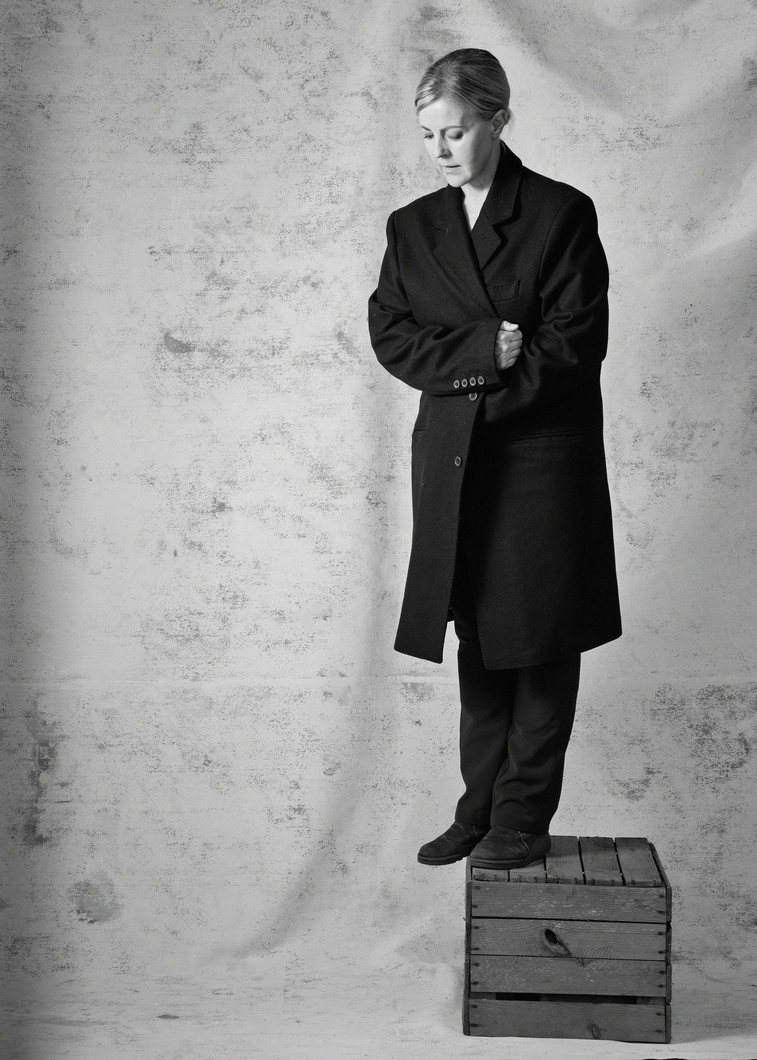 Black and white photo of a woman in an oversized dark coat standing on a wooden crate against a textured backdrop.