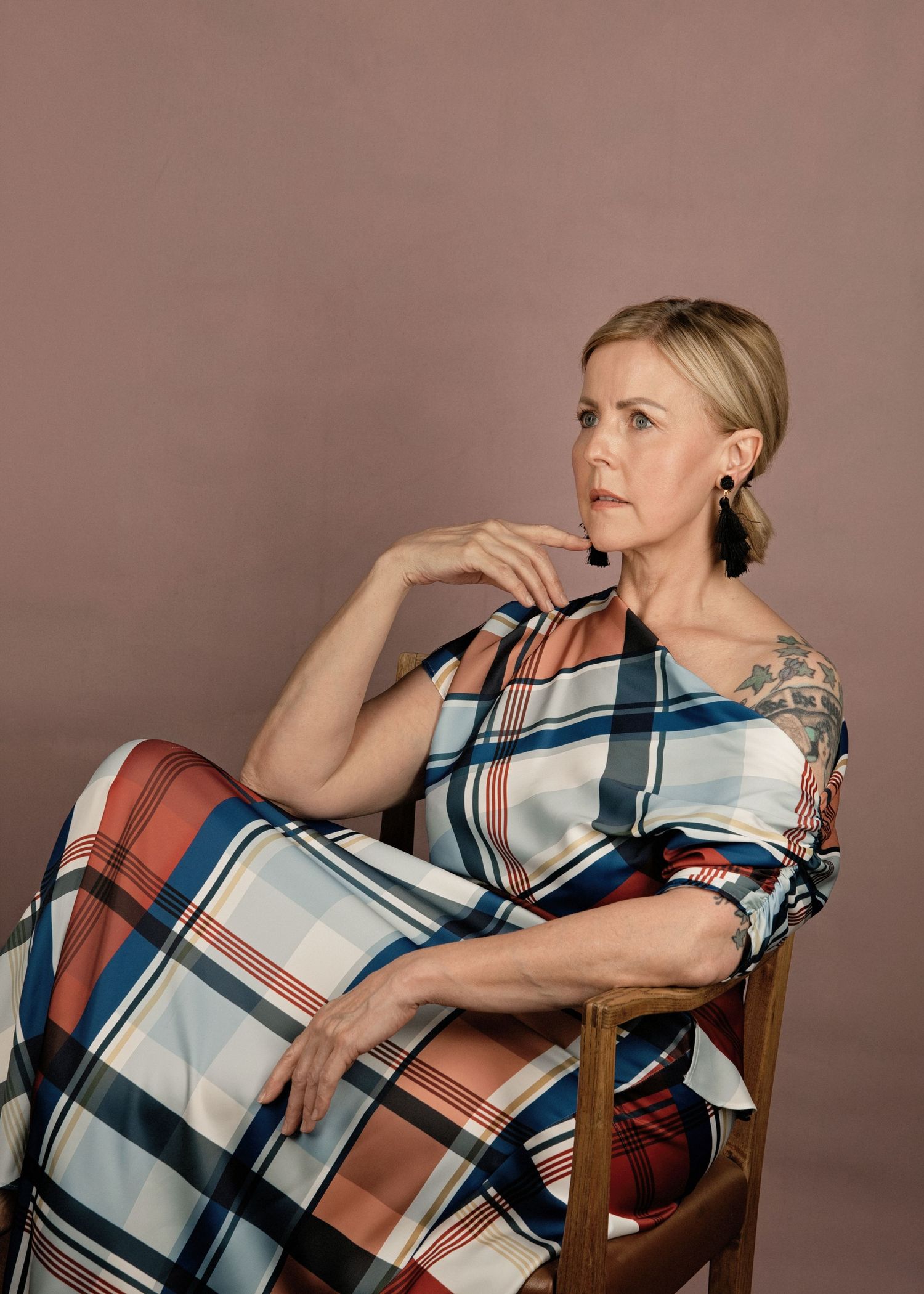 Woman in plaid dress seated in wooden chair, posing thoughtfully against a mauve studio background.