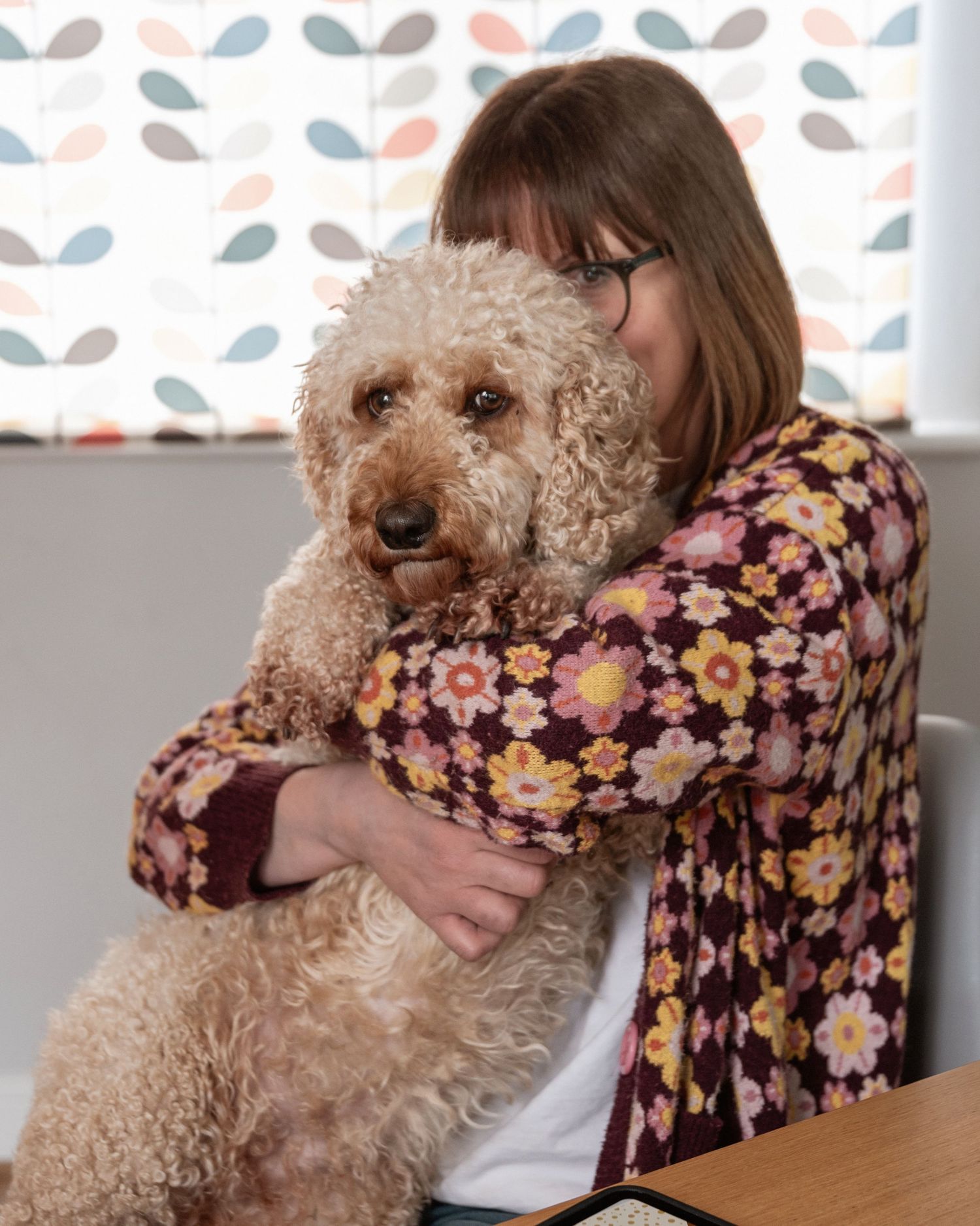 Woman in floral jacket hugging a fluffy cream Labradoodle dog indoors.